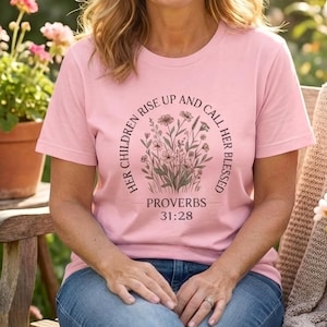 Puede incluir: Una camiseta rosa claro con el texto "HER CHILDREN RISE UP AND CALL HER BLESSED" y "PROVERBIOS 31:28" en un diseño floral circular. La persona lleva vaqueros azules y está sentada en un banco de madera en un jardín.