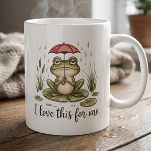 May include: White ceramic mug featuring a whimsical illustration of a frog under a red umbrella, sitting on a lily pad. The mug has the text "I love this for me" in a cursive font. The mug has a curved handle.