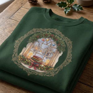 May include: A dark green crewneck sweatshirt featuring a circular graphic print of a window view. The illustration shows a stack of books, lit candles, and a festive wreath with ornaments and pine branches, all framed by an ornate gold border.