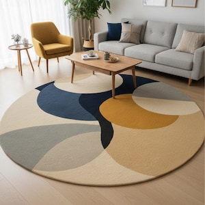 May include: A round area rug with a modern design. The rug features overlapping geometric shapes in shades of blue, gray, beige, and gold. The rug is placed on a hardwood floor in a living room setting.