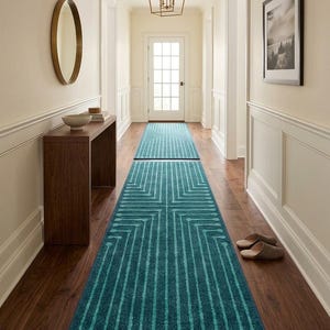 Op de afbeelding: Een lang, rechthoekig loperkleed met een diep blauwgroene achtergrond en een geometrisch patroon van lichtere blauwgroene lijnen die eindigen in afgeronde vormen. Het kleed ligt op een donkere houten vloer in een gang.