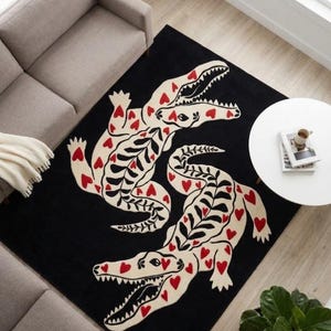 Puede incluir: Una alfombra rectangular con fondo negro que presenta dos grandes cocodrilos de color crema dispuestos en espejo. Los cocodrilos están decorados con pequeños corazones rojos y patrones negros en forma de hoja a lo largo de sus lomos.