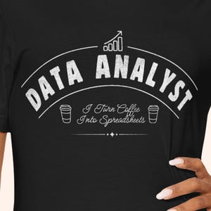 May include: Black t-shirt featuring a white graphic with the words "DATA ANALYST" in an arched design. The text "I Turn Coffee Into Spreadsheets" is below, with coffee cup illustrations. A chart icon is above.