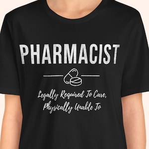 Puede incluir: Camiseta negra con la palabra "FARMACÉUTICO" en letras blancas. Debajo, un diseño de pastilla y el texto "Legalmente obligado a cuidar, físicamente incapaz de".