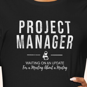 May include: Black t-shirt with white text that reads "PROJECT MANAGER" above a graphic and the text "WAITING ON AN UPDATE For a Meeting About a Meeting". The shirt is a crew neck.