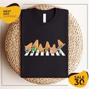 a black t-shirt with a graphic design of cartoon characters wearing sombreros, arranged in a crosswalk pattern. The t-shirt is displayed on a woven placemat, and there are two yellow sale signs in the image.