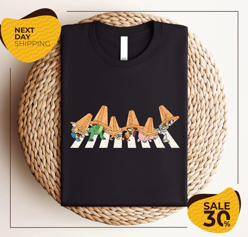 a black t-shirt with a graphic design of cartoon characters wearing sombreros, arranged in a crosswalk pattern. The t-shirt is displayed on a woven placemat, and there are two yellow sale signs in the image.