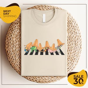 a beige t-shirt with a cartoon-style illustration of a crosswalk with cartoon characters wearing hats. The t-shirt is displayed on a woven basket, and there are two yellow sale signs in the image.