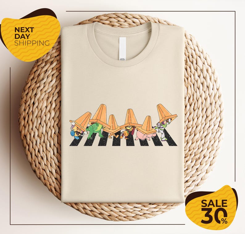 a beige t-shirt with a cartoon-style illustration of a crosswalk with cartoon characters wearing hats. The t-shirt is displayed on a woven basket, and there are two yellow sale signs in the image.