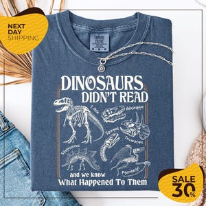 May include: A blue Comfort Colors t-shirt featuring a dinosaur-themed graphic. The graphic includes dinosaur skeletons and the text "DINOSAURS DIDN'T READ and we know What Happened To Them". A silver necklace is draped across the shirt.