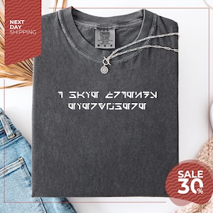 May include: Dark gray Comfort Colors t-shirt with white text in an alien language. A silver necklace with a pendant rests on the shirt. The shirt is folded, with a sale tag visible. Next day shipping is advertised.