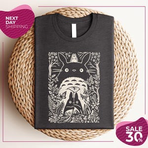 a gray t-shirt with a black and white graphic design depicting a cat surrounded by various plants and flowers. The t-shirt is placed on a woven placemat, and there are two pink circular sale signs in the image.