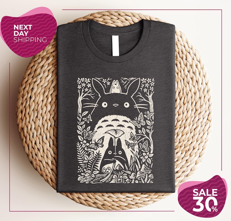 a gray t-shirt with a black and white graphic design depicting a cat surrounded by various plants and flowers. The t-shirt is placed on a woven placemat, and there are two pink circular sale signs in the image.