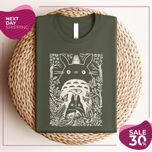 a green t-shirt with a white graphic design depicting a cat surrounded by various natural elements, such as leaves, flowers, and mushrooms. The t-shirt is displayed on a woven placemat, and there are two pink circular sale signs in the image, one indicating "Next Day Shipping" and the other offering a "Sale 30%".
