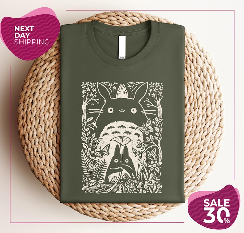 a green t-shirt with a white graphic design depicting a cat surrounded by various natural elements, such as leaves, flowers, and mushrooms. The t-shirt is displayed on a woven placemat, and there are two pink circular sale signs in the image, one indicating "Next Day Shipping" and the other offering a "Sale 30%".