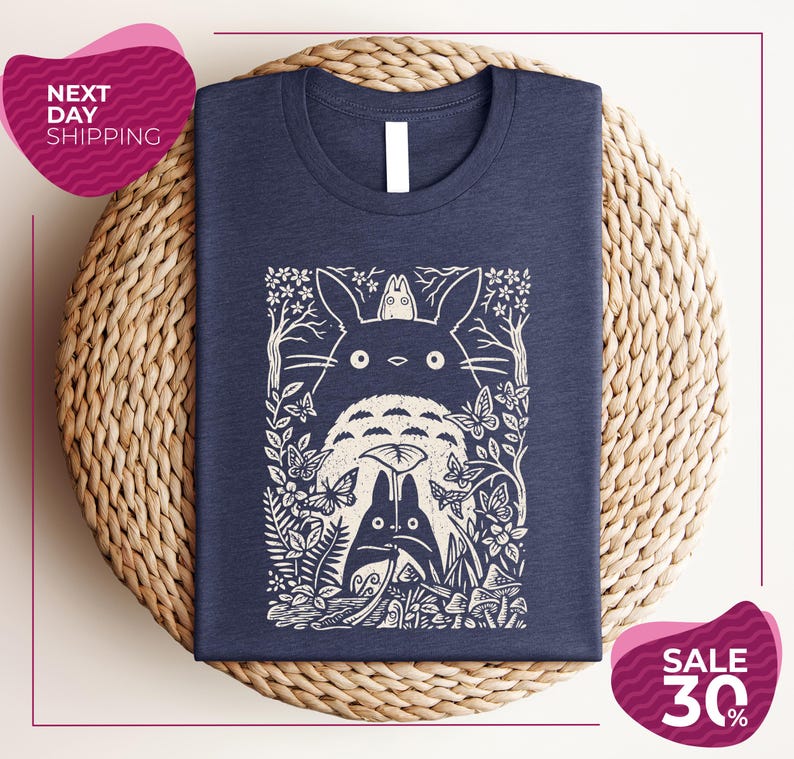 a blue t-shirt with a white graphic design depicting a cat surrounded by various natural elements, such as flowers and leaves. The t-shirt is placed on a woven placemat, and there are two pink circular signs in the image. One sign advertises "Next Day Shipping" and the other sign advertises a "SALE 30%".