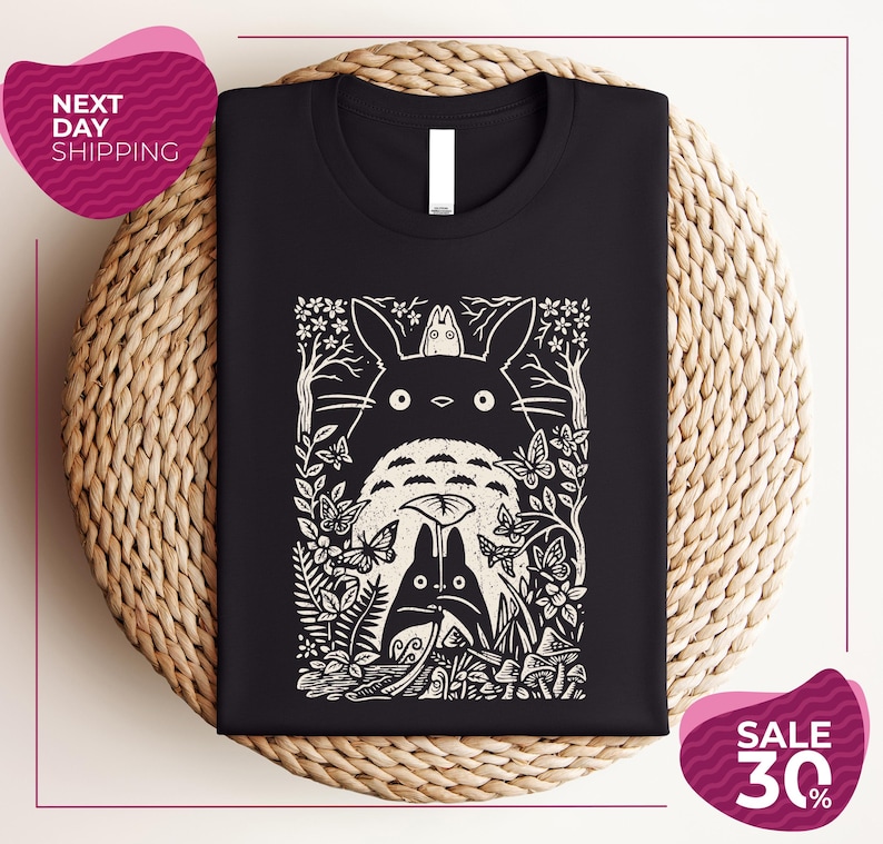 a black t-shirt with a white graphic design depicting a cat surrounded by various elements such as flowers, leaves, and mushrooms. The t-shirt is placed on a woven placemat, and there are two pink circular sale signs in the image.