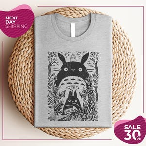 a gray t-shirt with a black and white graphic design depicting a cat surrounded by various plants and flowers. The t-shirt is placed on a woven placemat, and there are two pink circular sale signs in the image.