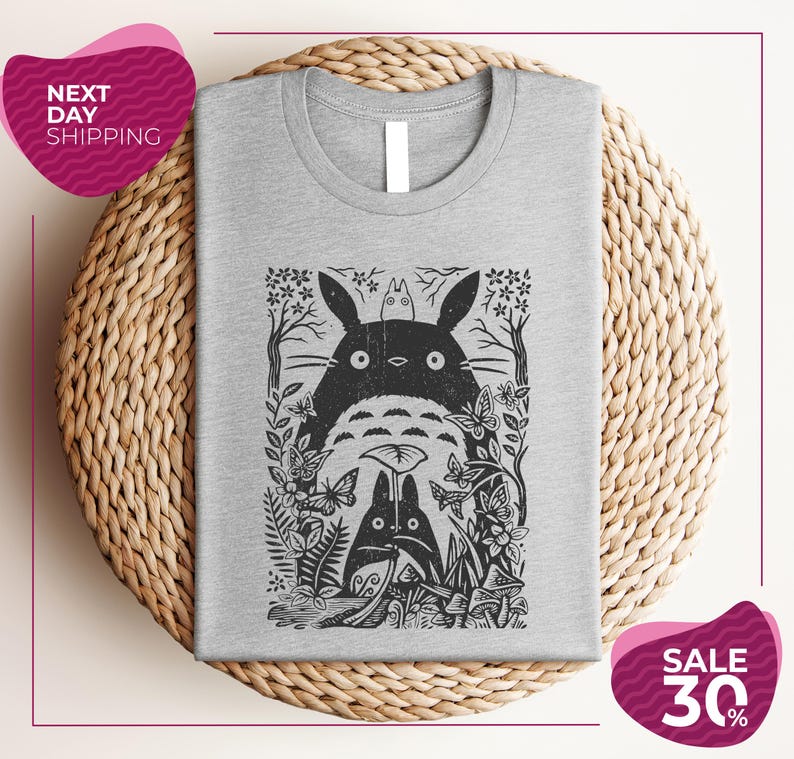a gray t-shirt with a black and white graphic design depicting a cat surrounded by various plants and flowers. The t-shirt is placed on a woven placemat, and there are two pink circular sale signs in the image.