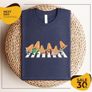 a blue t-shirt with a graphic design of cartoon characters crossing a crosswalk. The t-shirt is displayed on a woven basket, and there are two yellow price tags in the image.