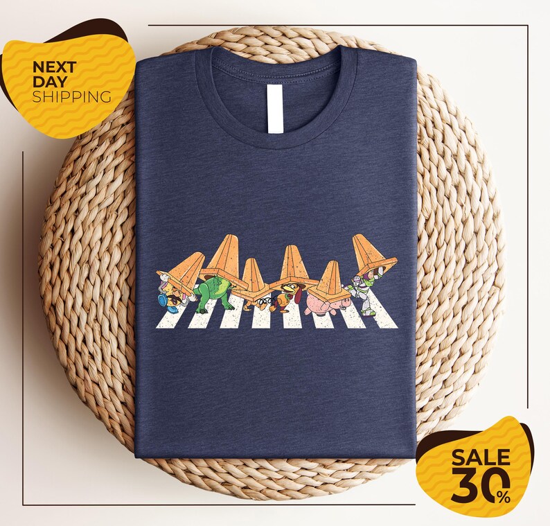 a blue t-shirt with a graphic design of cartoon characters crossing a crosswalk. The t-shirt is displayed on a woven basket, and there are two yellow price tags in the image.
