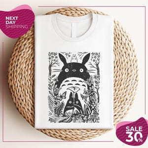 a white t-shirt with a black and white graphic design depicting a cat surrounded by various plants and flowers. The t-shirt is placed on a woven placemat, and there are two pink circular sale signs in the image.