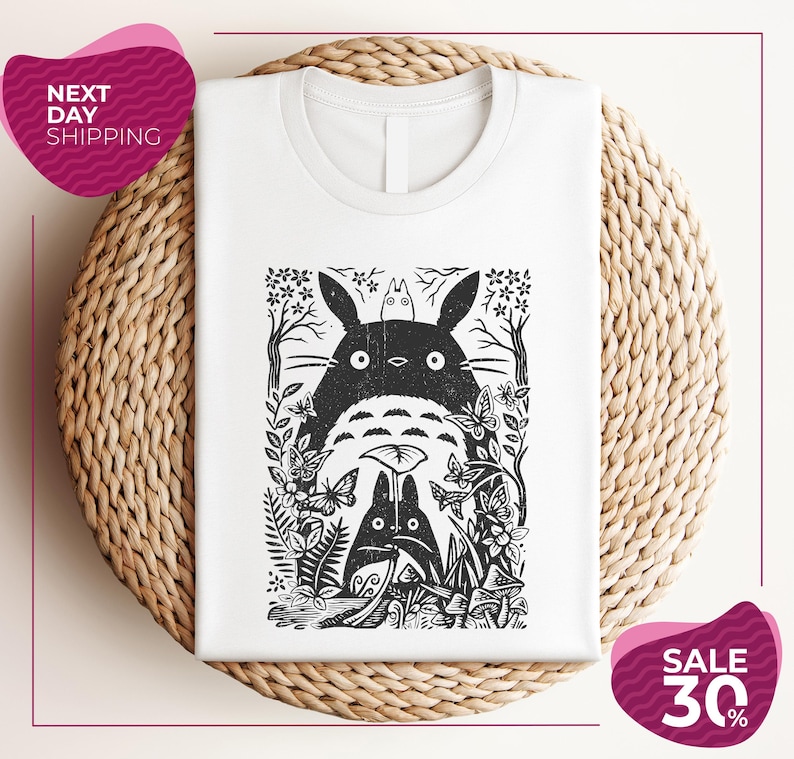 a white t-shirt with a black and white graphic design depicting a cat surrounded by various plants and flowers. The t-shirt is placed on a woven placemat, and there are two pink circular sale signs in the image.