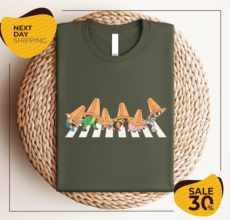 a green t-shirt with a graphic design of cartoon characters wearing orange hats, arranged in a crosswalk pattern. The t-shirt is displayed on a woven basket, and there are two yellow sale signs in the image.