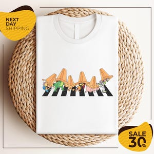 a white t-shirt with a colorful graphic design depicting a group of cartoon characters wearing hats and crossing a crosswalk. The t-shirt is displayed on a woven placemat, and there are two yellow sale signs in the image.
