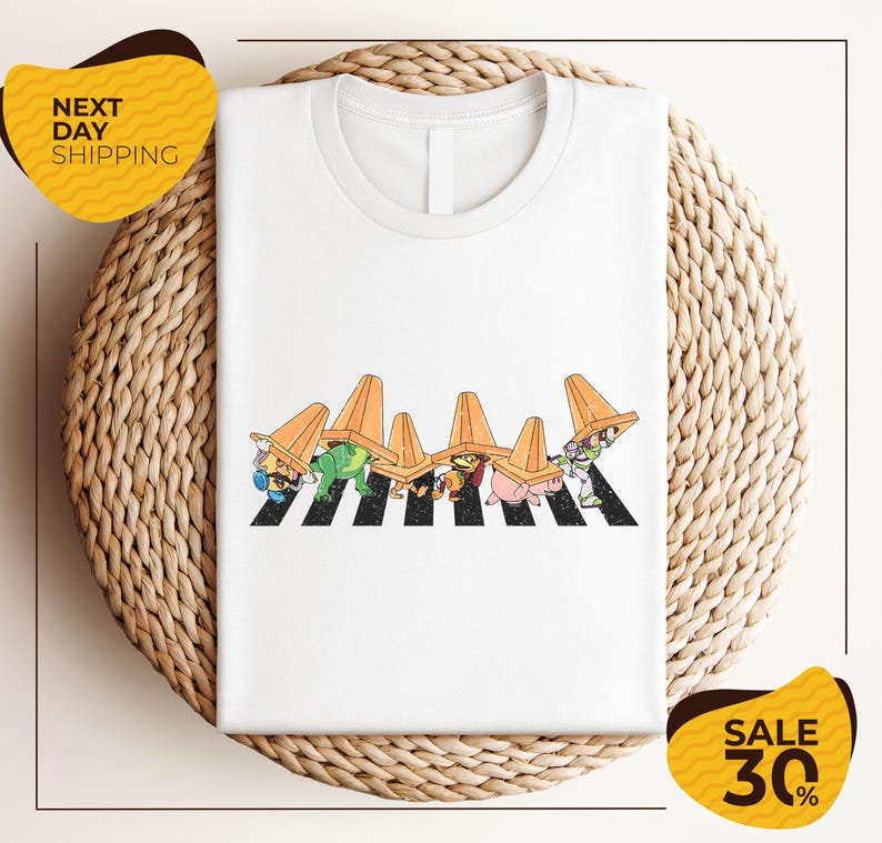 a white t-shirt with a colorful graphic design depicting a group of cartoon characters wearing hats and crossing a crosswalk. The t-shirt is displayed on a woven placemat, and there are two yellow sale signs in the image.