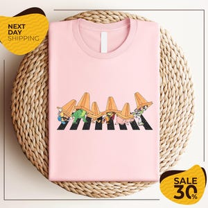 A pink t-shirt with a cartoon illustration of a group of people wearing hats, arranged in a crosswalk pattern. The t-shirt is displayed on a woven placemat, and there are yellow sale signs indicating a 30% discount.