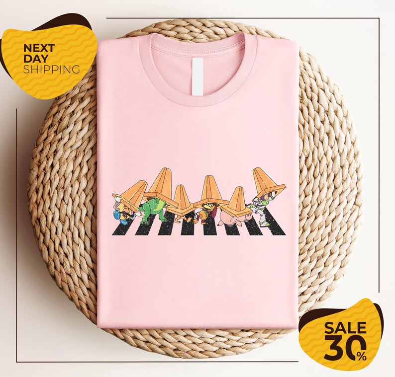 A pink t-shirt with a cartoon illustration of a group of people wearing hats, arranged in a crosswalk pattern. The t-shirt is displayed on a woven placemat, and there are yellow sale signs indicating a 30% discount.