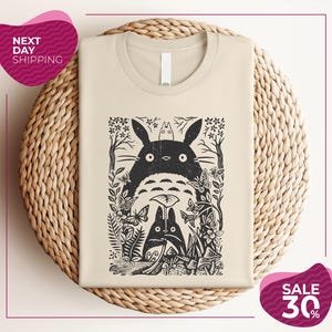 a beige t-shirt with a black and white graphic design depicting a cat surrounded by various plants and flowers. The t-shirt is placed on a woven basket, and there are two pink circular sale signs in the image.