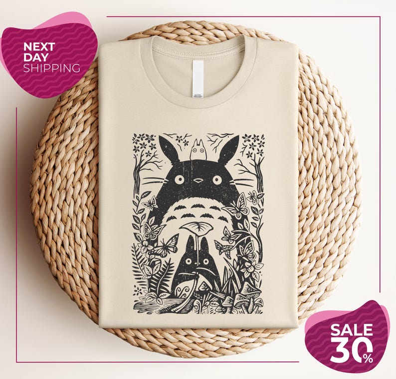 a beige t-shirt with a black and white graphic design depicting a cat surrounded by various plants and flowers. The t-shirt is placed on a woven basket, and there are two pink circular sale signs in the image.