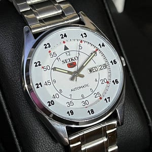 May include: A silver-toned Seiko automatic wristwatch with a white face. The watch features black numerals, red accents, and a day-date display. The watchband is made of silver-toned metal links.