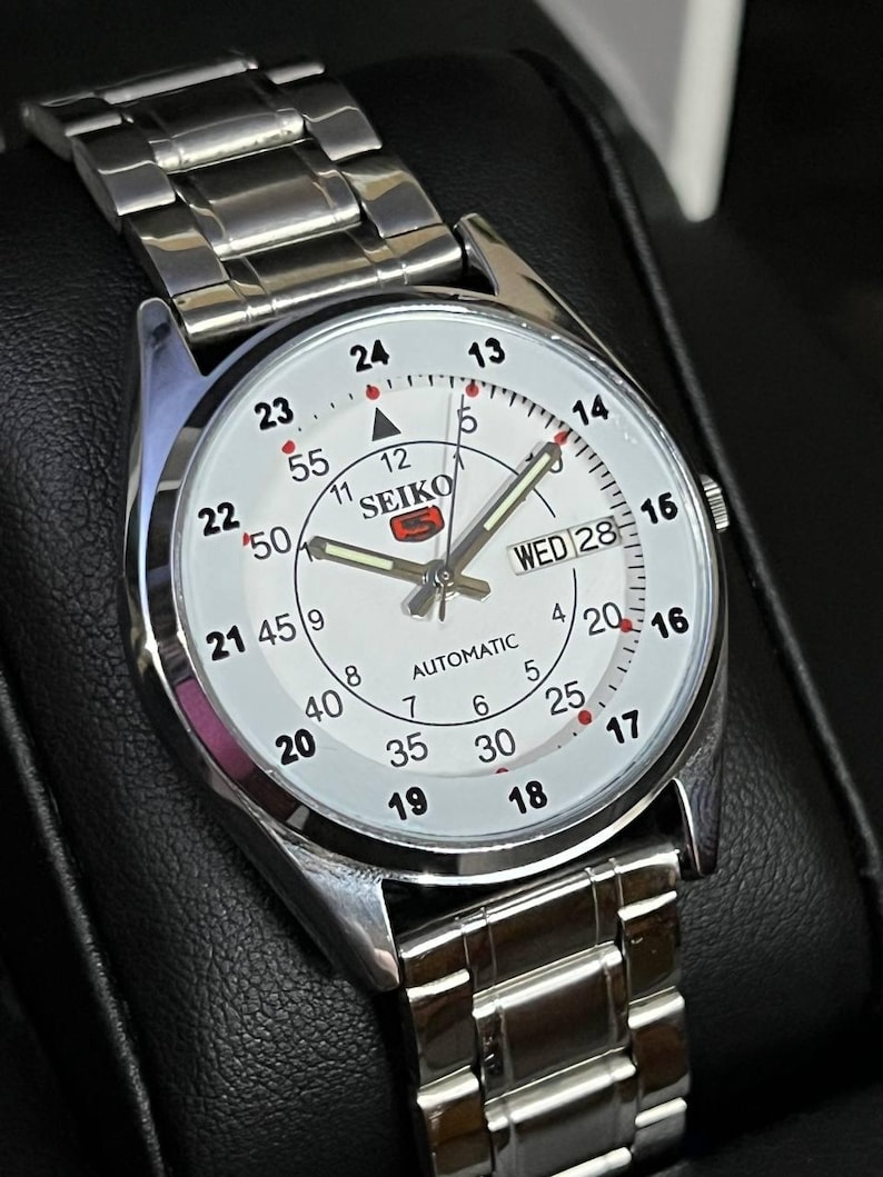 May include: A silver-toned Seiko automatic wristwatch with a white face. The watch features black numerals, red accents, and a day-date display. The watchband is made of silver-toned metal links.