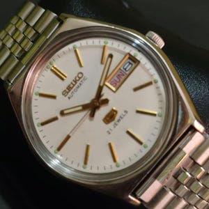 Puede incluir: Un reloj de pulsera autom&aacute;tico Seiko plateado con esfera blanca. El reloj presenta agujas e indicadores dorados, una pantalla de d&iacute;a y fecha, y una correa de metal plateado. La esfera del reloj indica "Seiko Automatic" y "21 Jewels".