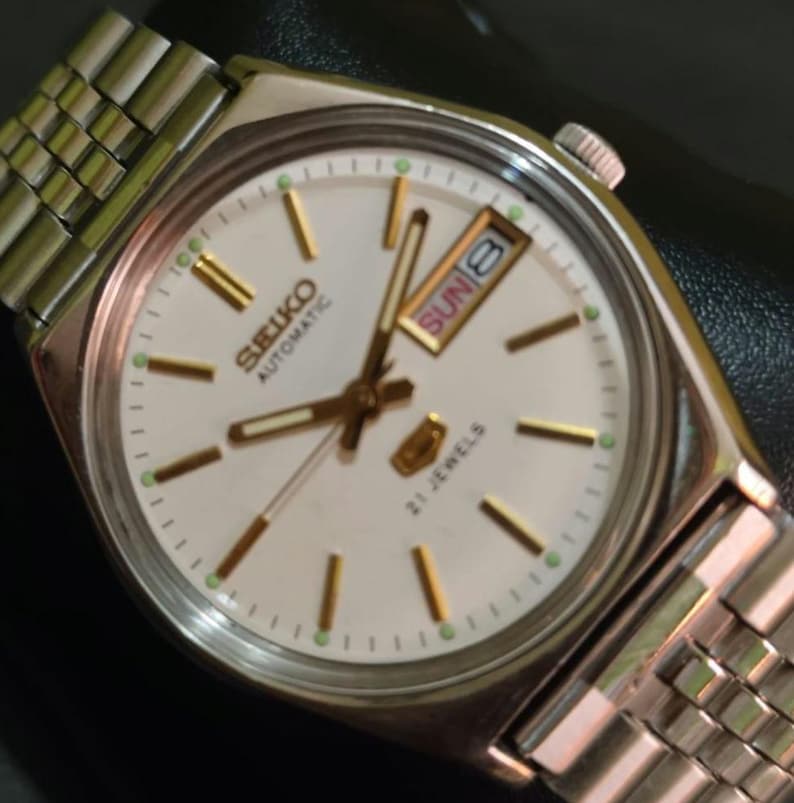 Puede incluir: Un reloj de pulsera autom&aacute;tico Seiko plateado con esfera blanca. El reloj presenta agujas e indicadores dorados, una pantalla de d&iacute;a y fecha, y una correa de metal plateado. La esfera del reloj indica "Seiko Automatic" y "21 Jewels".