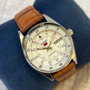 Puede incluir: Un reloj de pulsera automático Seiko vintage con esfera blanca, caja plateada y correa de cuero marrón. La esfera muestra el día y la fecha, con un dial de 24 horas. Fabricado en Japón.