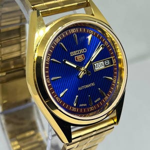 May include: A gold-coloured Seiko 5 automatic wristwatch with a blue dial. The watch face displays the day and date, with gold hands and markers. The watch band is also gold-coloured.