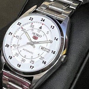 May include: Silver-toned Seiko automatic wristwatch with a white face. The watch features black numerals, a day-date display, and a silver metal band. The watch face displays the word "SEIKO".