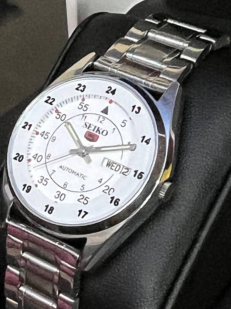 May include: Silver-toned Seiko automatic wristwatch with a white face. The watch features black numerals, a day-date display, and a silver metal band. The watch face displays the word "SEIKO".