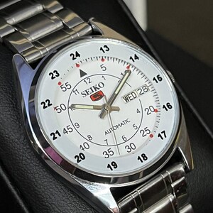 May include: A silver-toned Seiko automatic wristwatch with a white face. The watch features a day and date display, along with a silver band. The watch face displays the time in both 12-hour and 24-hour formats.