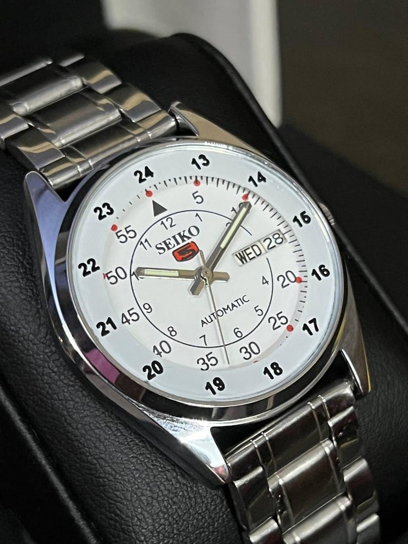 May include: A silver-toned Seiko automatic wristwatch with a white face. The watch features a day and date display, along with a silver band. The watch face displays the time in both 12-hour and 24-hour formats.
