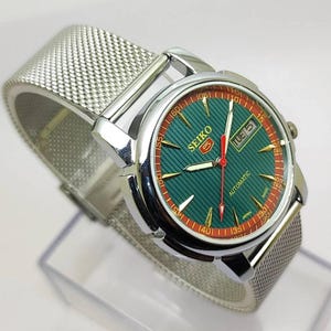 May include: A silver-toned Seiko automatic wristwatch with a green dial and a mesh band. The watch face features gold-toned hour markers, a day-date display, and red accents. The watch is displayed on a clear stand.