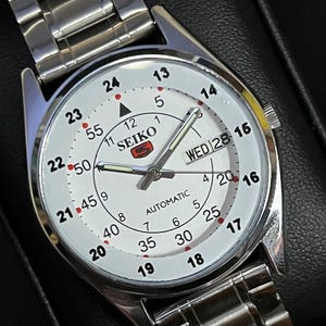 May include: A silver-toned Seiko automatic wristwatch with a white face. The watch features black numerals, a day-date display, and a silver band. The watch face displays the brand name and the word "AUTOMATIC".