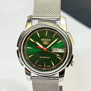 May include: A silver-toned Seiko 5 automatic watch with a green dial and a mesh band. The watch face features gold-colored hands and markers, a day-date display, and the text "SEIKO 5 AUTOMATIC JAPAN MADE".