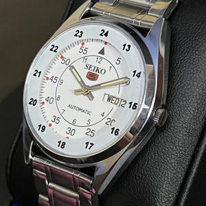 May include: A silver-toned Seiko automatic wristwatch with a white face. The watch features black numerals, a day-date display, and a silver-toned band. The watch face displays the brand name and the word "automatic".
