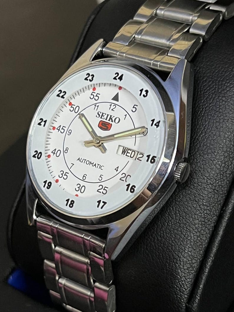 May include: A silver-toned Seiko automatic wristwatch with a white face. The watch features black numerals, a day-date display, and a silver-toned band. The watch face displays the brand name and the word "automatic".