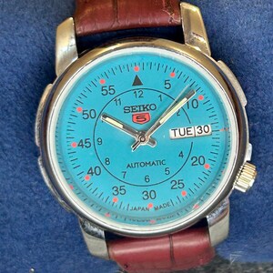 May include: A Seiko 5 automatic wristwatch with a bright turquoise dial, silver-tone hands, and a day-date window at the three o'clock position. The watch features a brown leather strap with a textured finish and a stainless steel case.